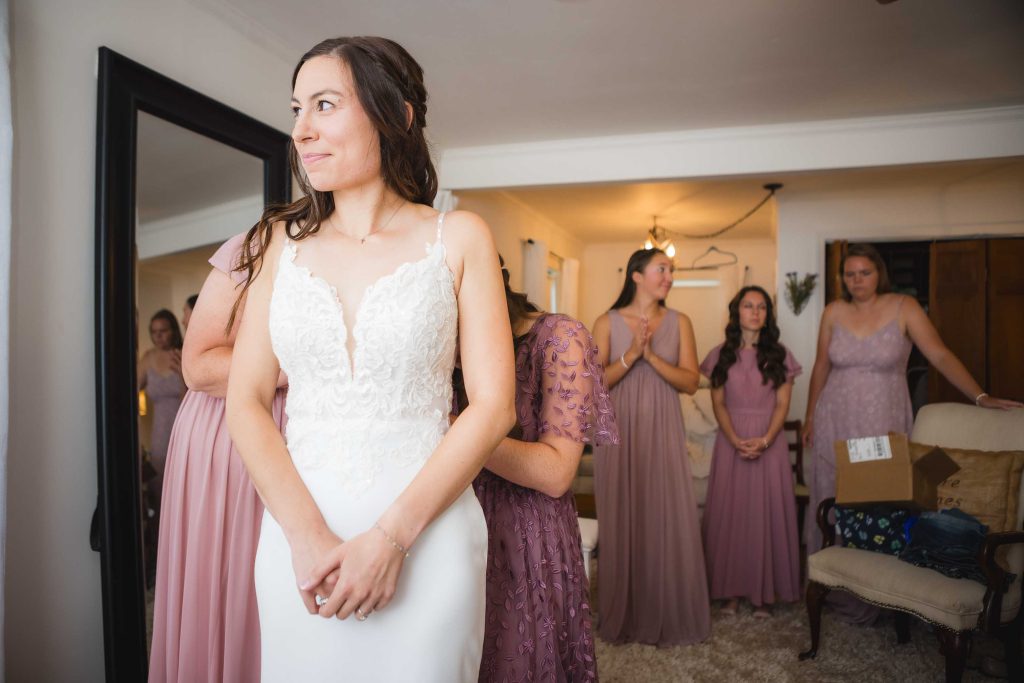 A candid wedding preparation moment with the bride and her bridesmaids standing in front of a mirror.