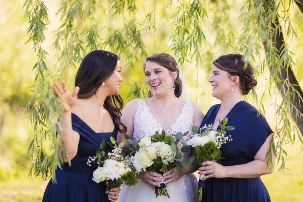 Three bridesmaids laughing in front of a willow tree at a wedding.