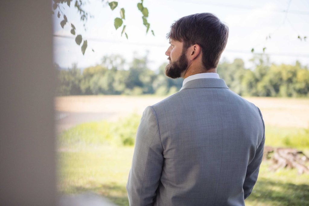 A groom in a suit is looking out of a window during wedding preparation.
