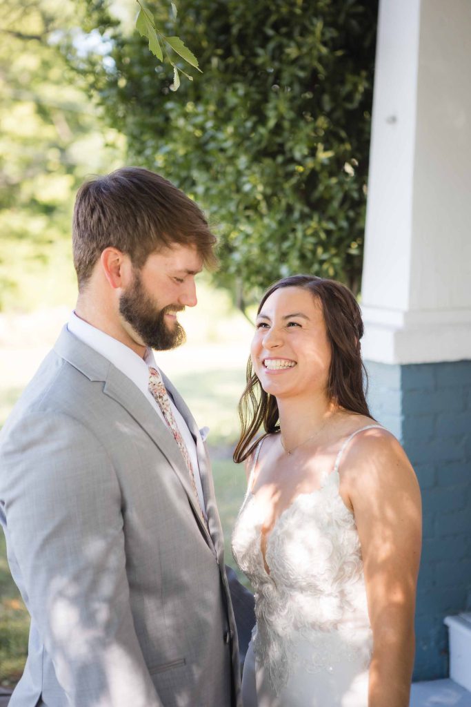 A candid wedding couple smiling at each other in front of a house.