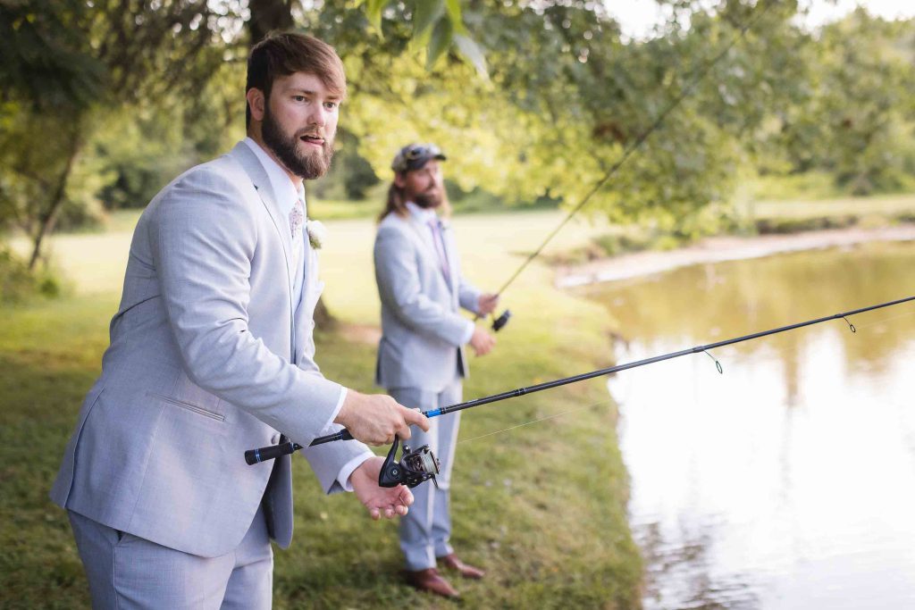 Two groomsmen posing for a wedding portrait, holding fishing rods in front of a pond.