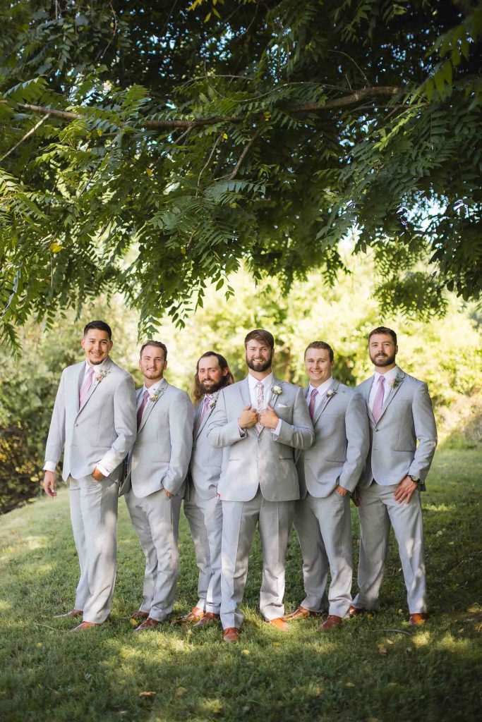 A group of groomsmen posing for a wedding portrait in front of a tree.