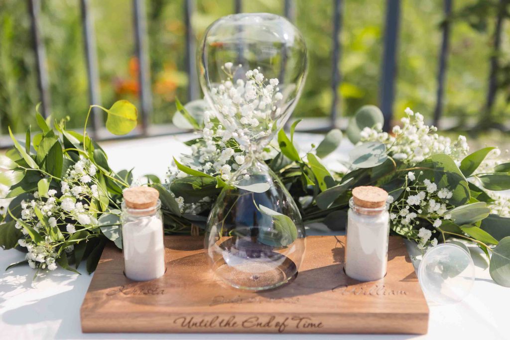 A glass hourglass on a wooden table with detailed flowers and greenery, perfect for a wedding.