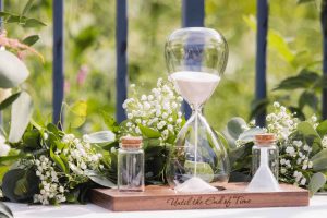 A sand hourglass sits on a table next to some flowers, adding a touch of wedding details.