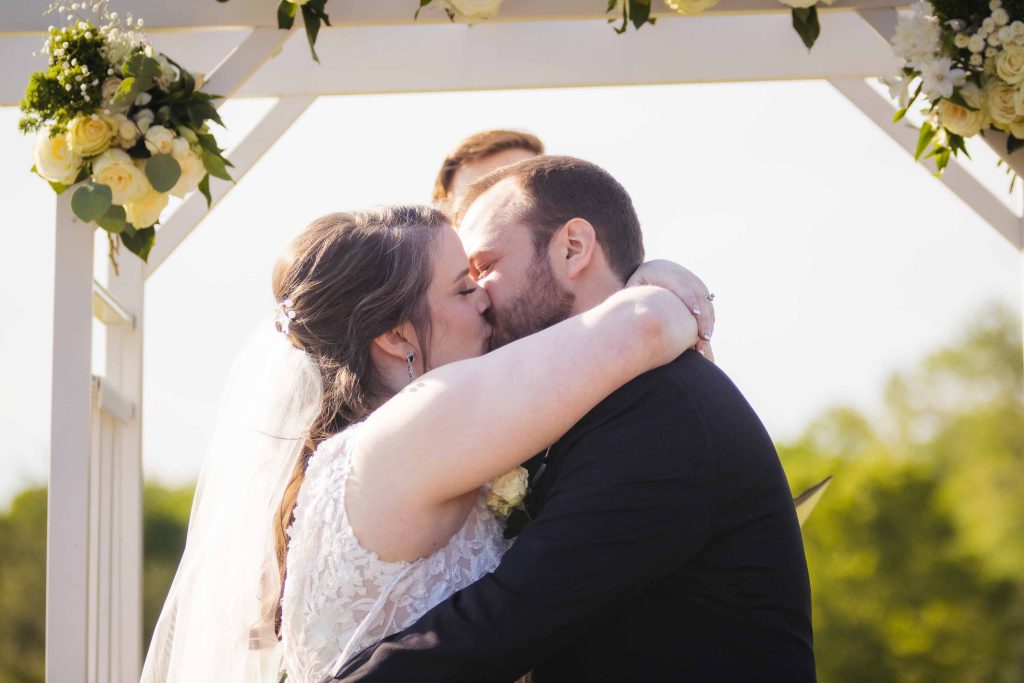 A bride and groom share a kiss during their wedding ceremony under a beautiful arch.