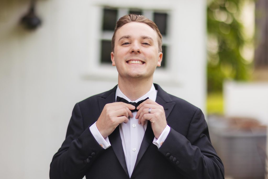 A man in a tuxedo adjusting his bow tie for a wedding portrait.