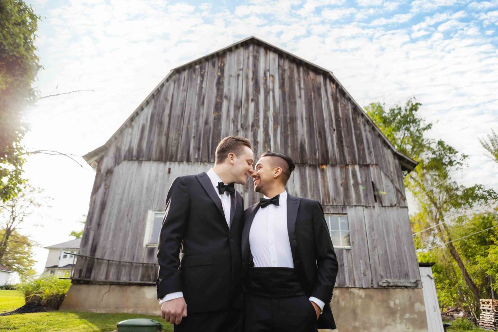 Two grooms share a wedding kiss in front of a barn.