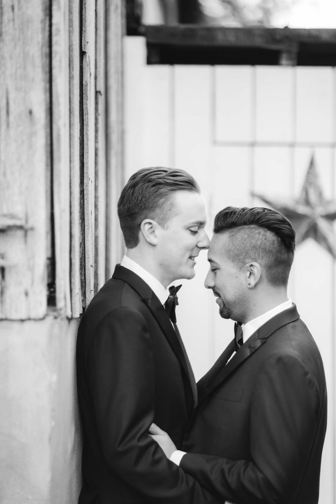 Two men in tuxedos hugging in front of a barn, capturing a wedding portrait.