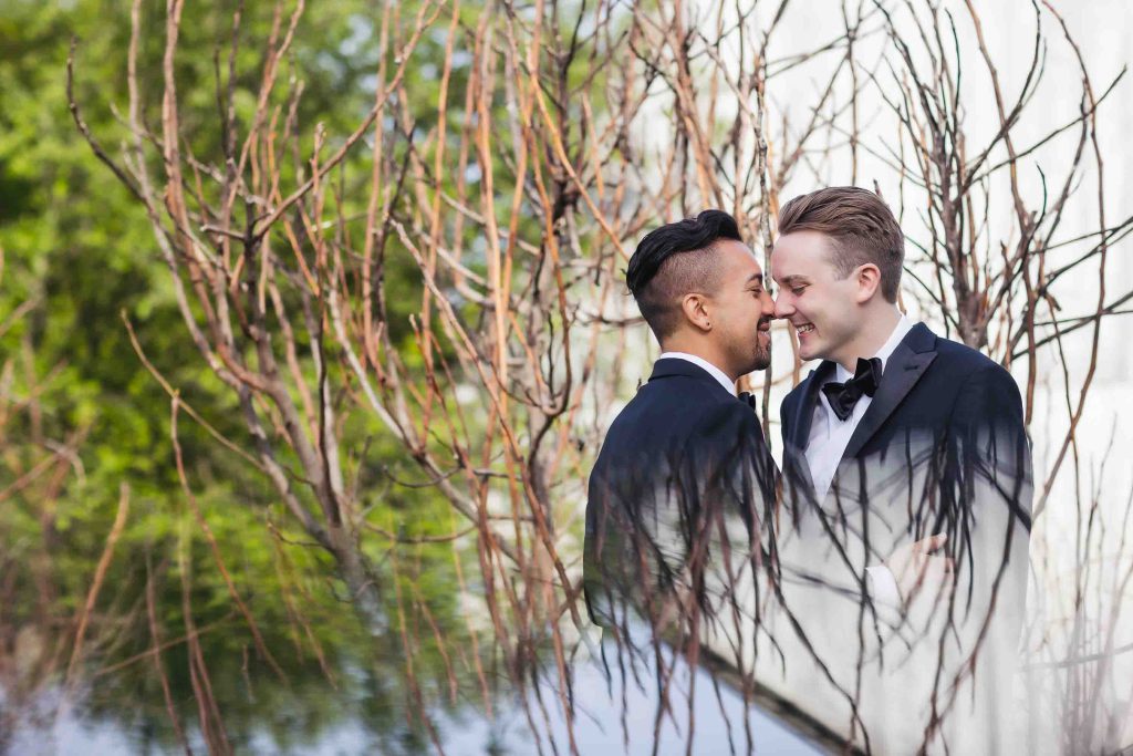 Wedding portrait of two men in tuxedos kissing in front of a tree.