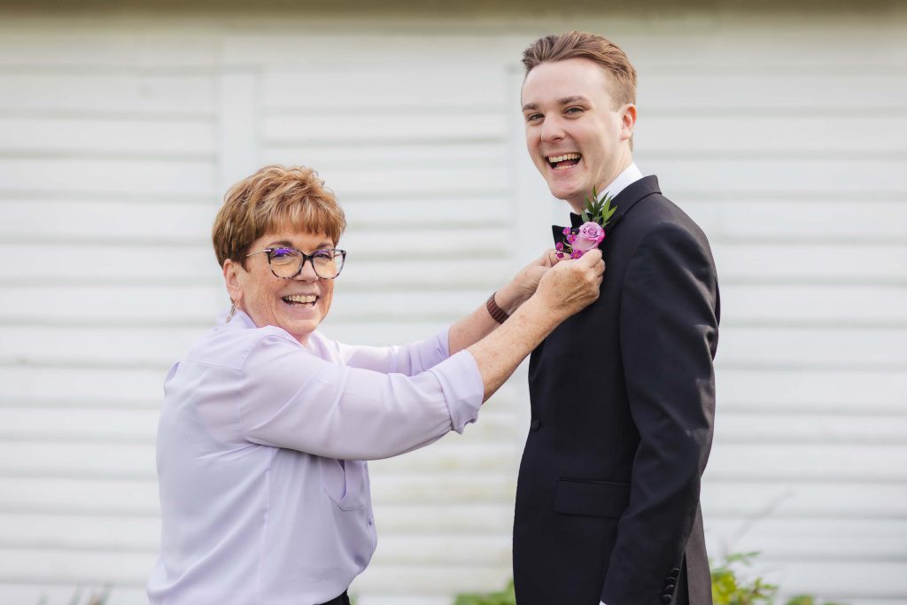 A candid wedding preparation moment as a woman puts a boutonniere on a man in a tuxedo.