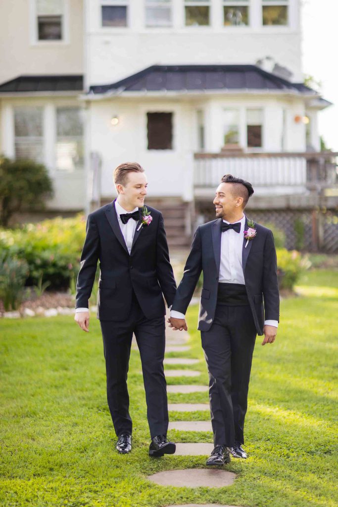 Two men in tuxedos holding hands in front of a house for a wedding portrait.