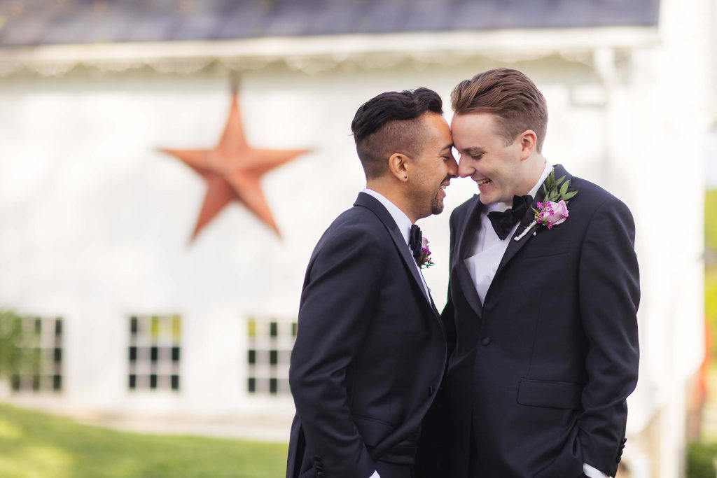 Two men in tuxedos are smiling in front of a barn for a wedding portrait.
