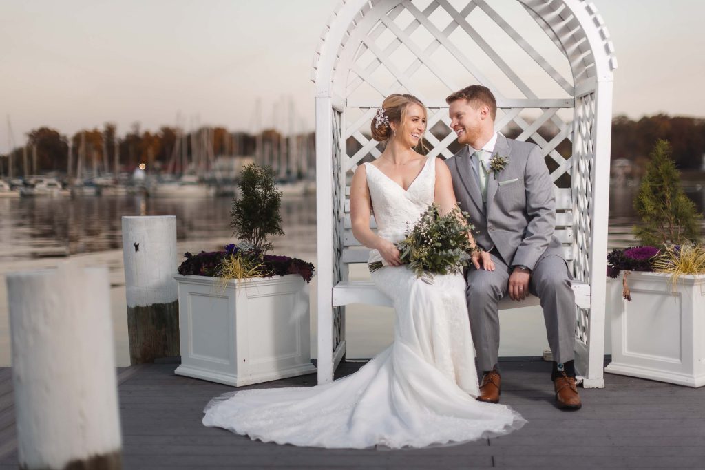 A wedding portrait of a bride and groom on a wooden bench by a dock.