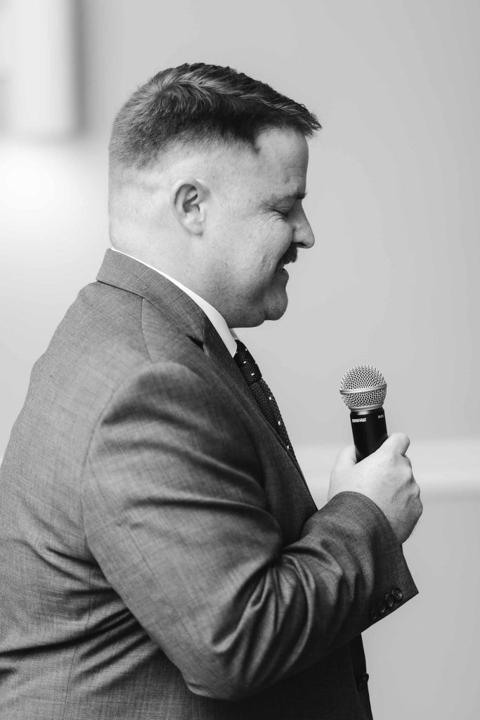 A man in a suit speaking at a wedding reception.