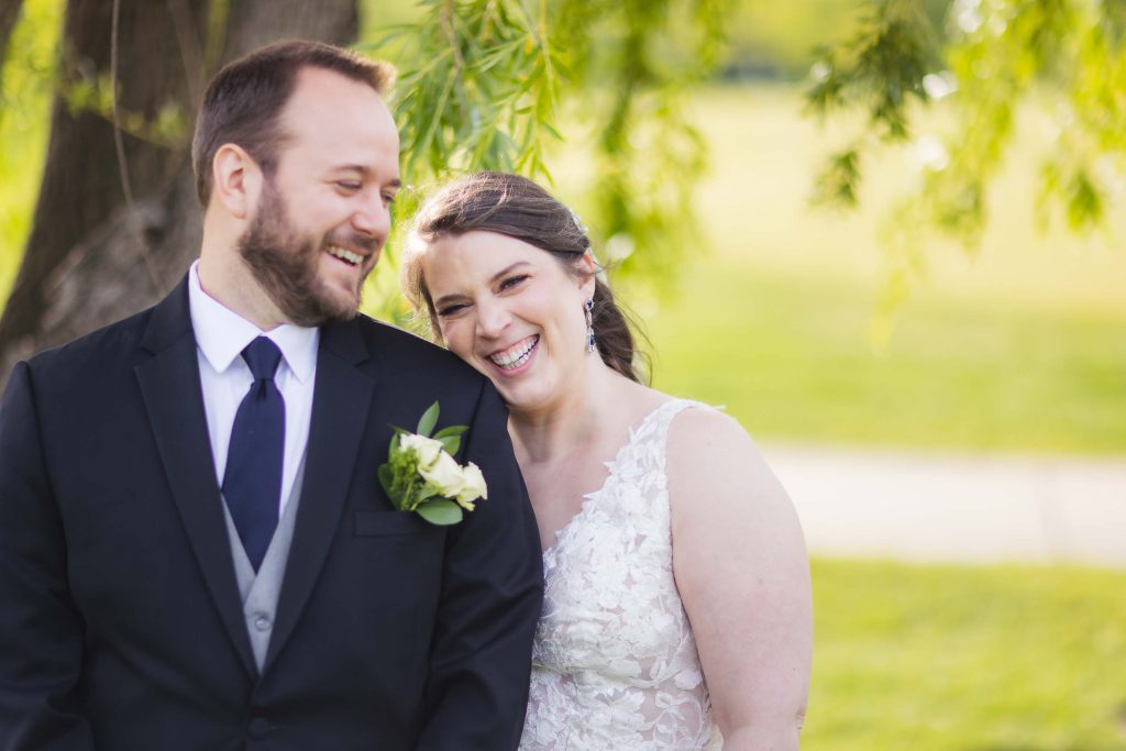 A wedding portrait of a smiling bride and groom in front of a tree.