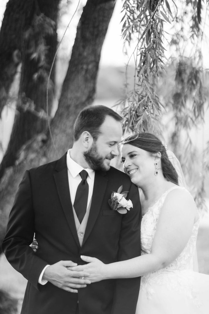 A black and white portrait of a bride and groom in front of a willow tree.