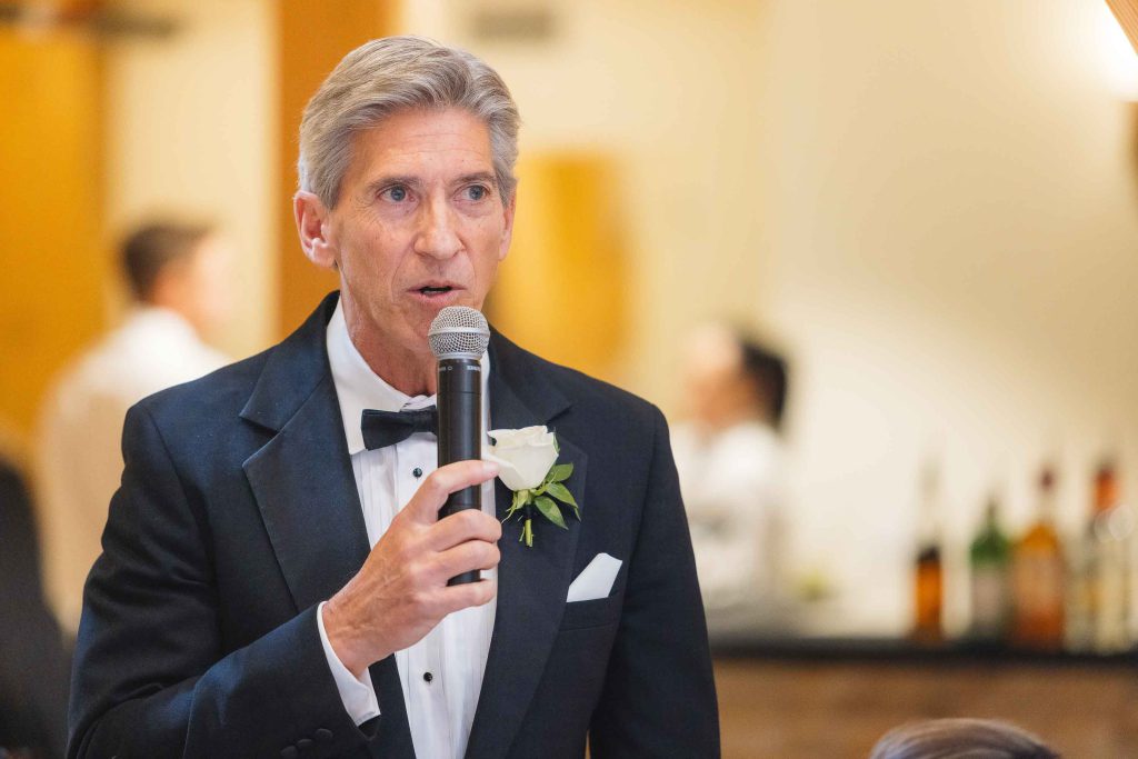 A man in a tuxedo speaking at a wedding reception.