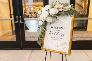 A detailed welcome sign with flowers and greenery in front of a wedding door.