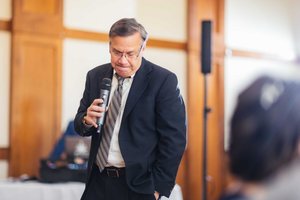 A man in a suit speaking into a microphone at a wedding reception.