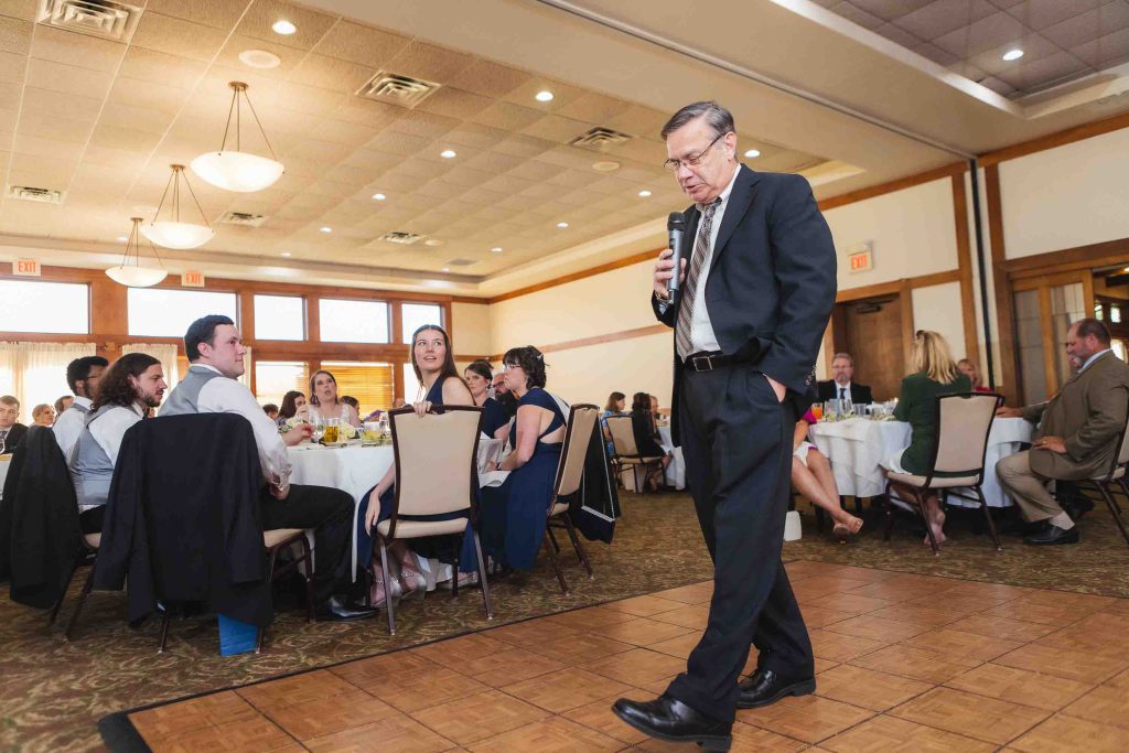 A man in a suit speaking into a microphone at a wedding reception.