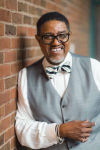 A man in a gray vest and bow tie affiliated with Hesson Entertainment leaning against a brick wall.