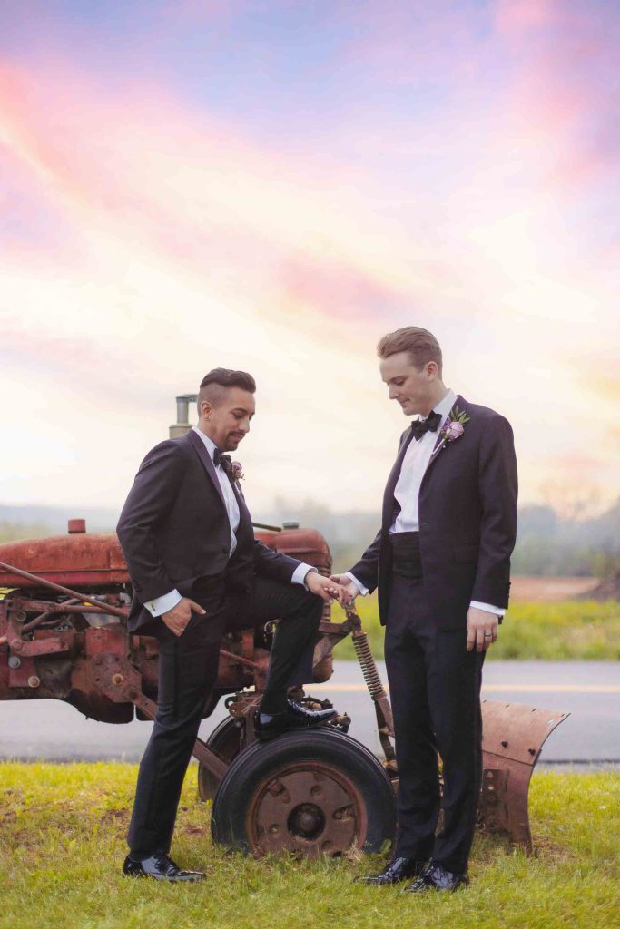 Two men in tuxedos posing next to a tractor at a wedding.