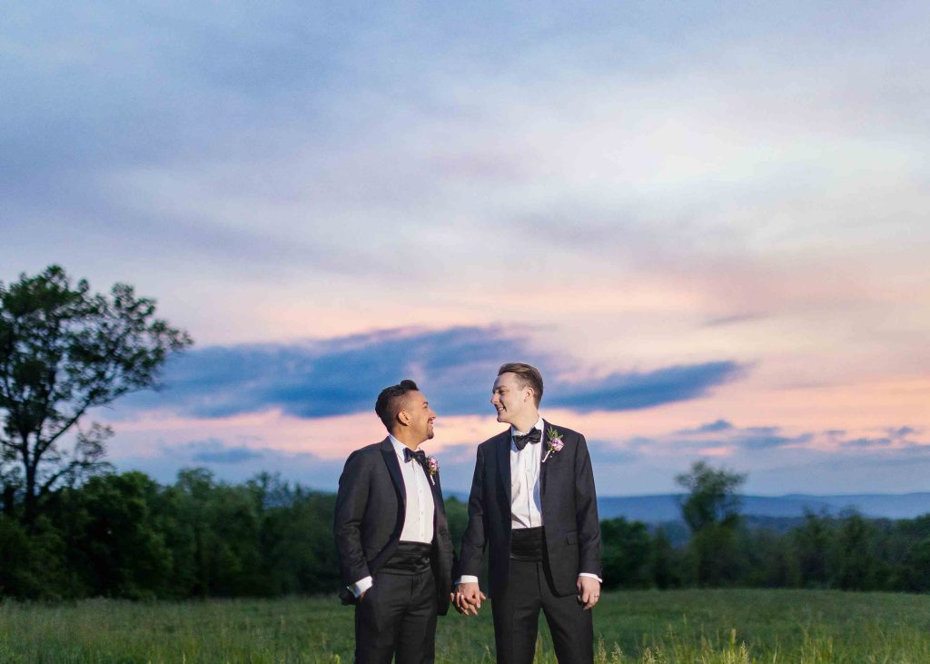 Two grooms in tuxedos posing for a wedding portrait in a field at sunset.