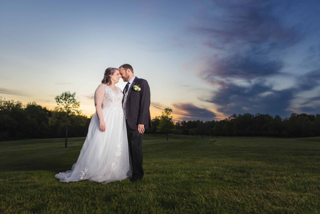 A wedding portrait of a bride and groom kissing in a field at sunset.