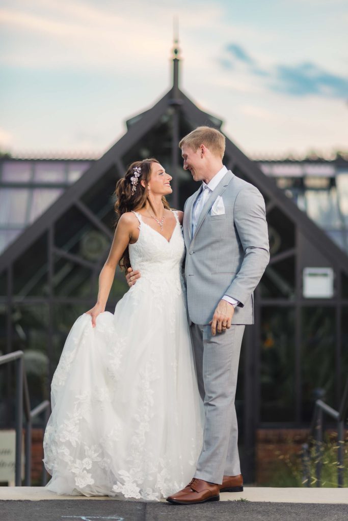 A wedding portrait of a bride and groom standing in front of a greenhouse.
