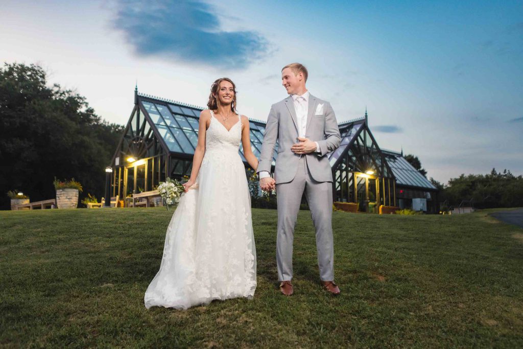 A wedding portrait of a bride and groom standing in front of a greenhouse at dusk.
