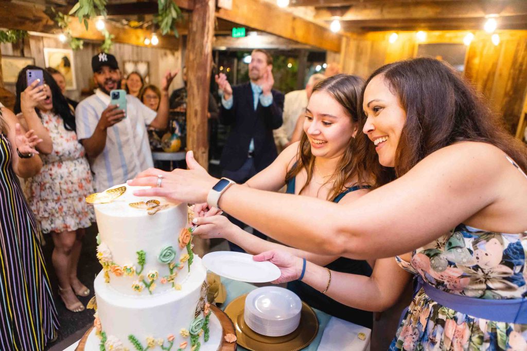 Two women cutting a birthday party cake in front of a crowd.