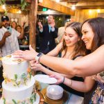 Two women cutting a birthday party cake in front of a crowd.