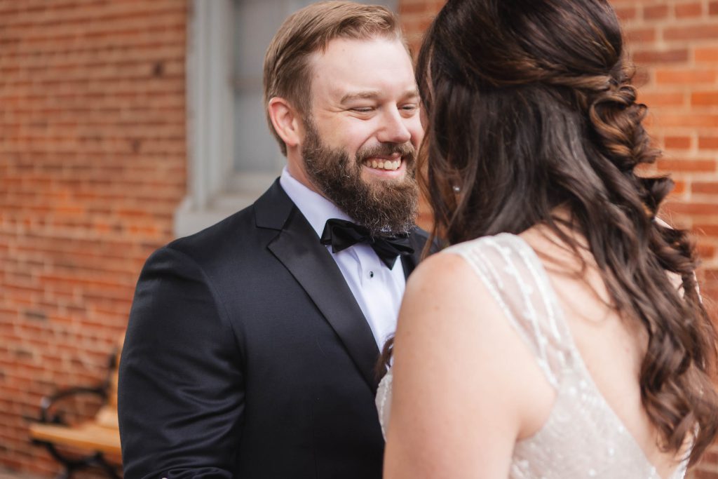 A candid bride and groom smiling at each other in front of the historic Savage Mill building during their wedding.