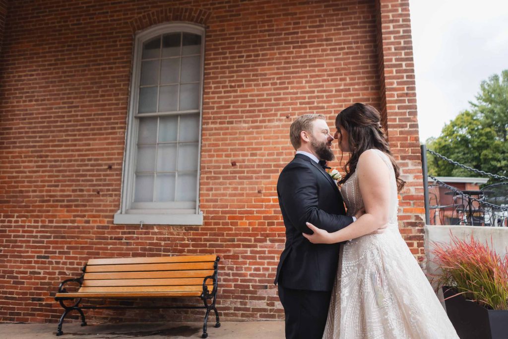 A candid moment captured at a wedding as the bride and groom share a passionate kiss in front of the historic Savage Mill brick building.
