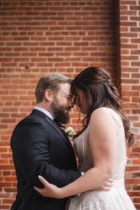 A candid bride and groom embrace in front of a brick wall at Savage Mill on their wedding day.