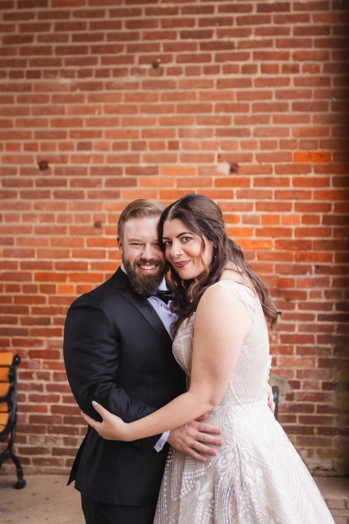 A candid wedding moment captured at Savage Mill, with the bride and groom embracing in front of a brick wall.