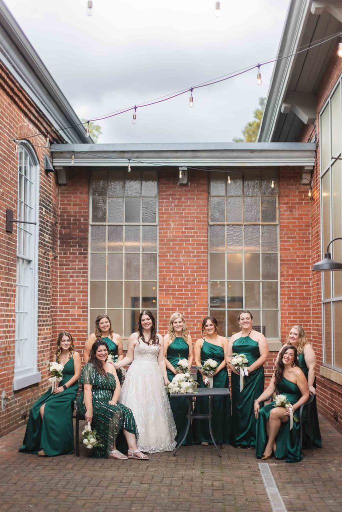 A group of bridesmaids in emerald dresses pose in front of a Savage Mill building.