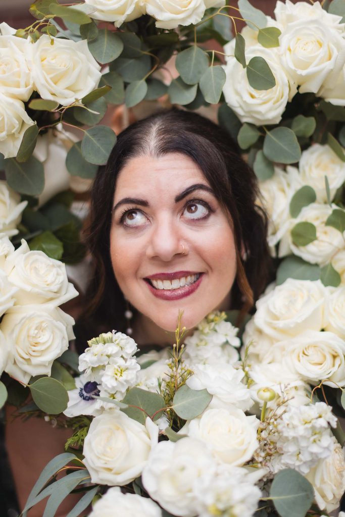 A wedding portrait of a bride standing gracefully in front of a wreath of white roses at Savage Mill.