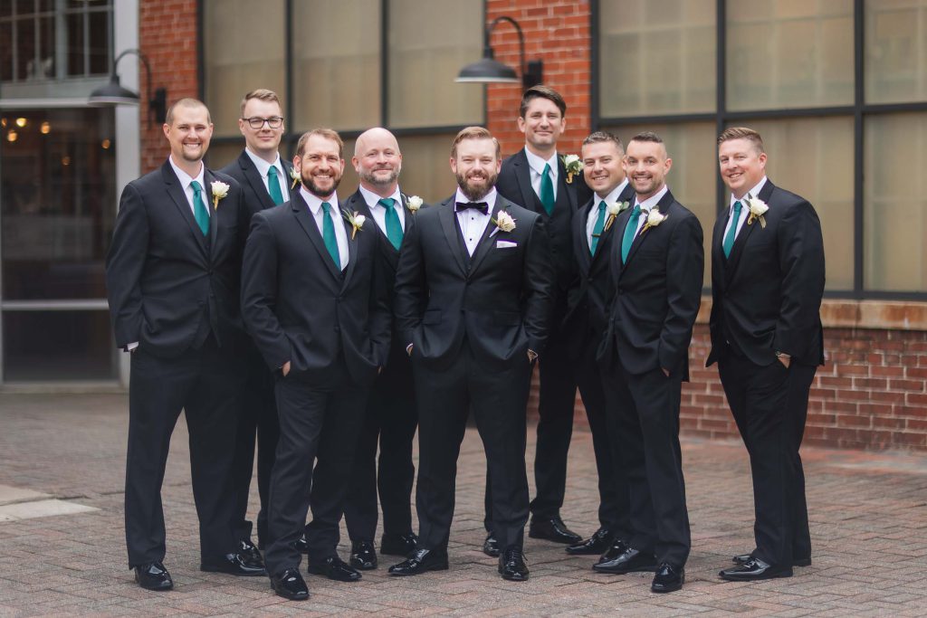 Groomsmen in black tuxedos standing in front of Savage Mill, a brick building, for a wedding portrait.