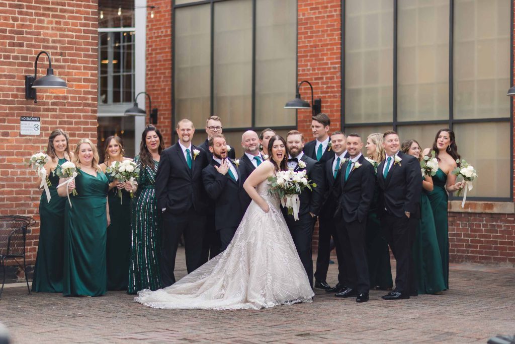 A group of bridesmaids and groomsmen pose for a wedding portrait in front of the Savage Mill's brick building.
