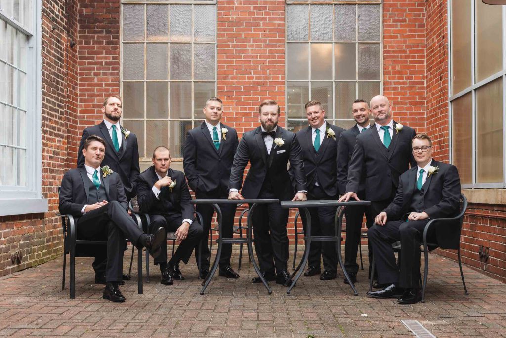 A group of groomsmen posing in front of a brick building for a wedding portrait at Savage Mill.