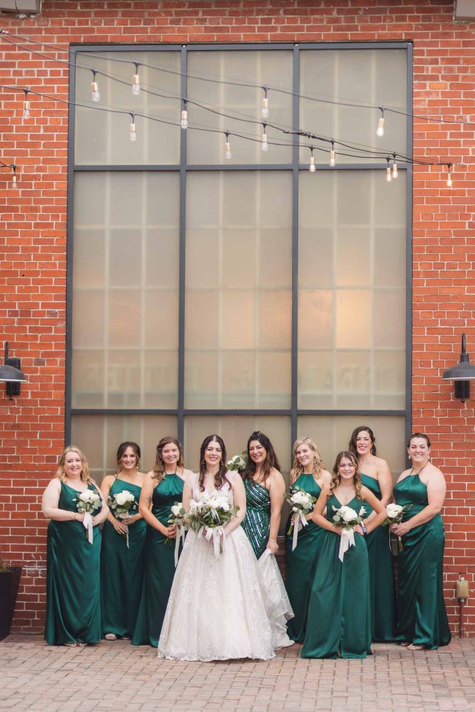 Bridesmaids in emerald dresses posing for a wedding portrait in front of the iconic brick building at Savage Mill.