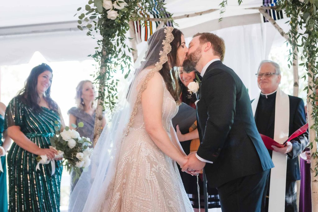 A bride and groom share a passionate kiss under a tent during their wedding ceremony at Savage Mill.