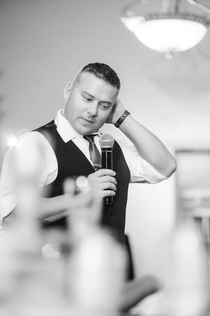 A man in a vest is holding a microphone at a wedding reception at Savage Mill.
