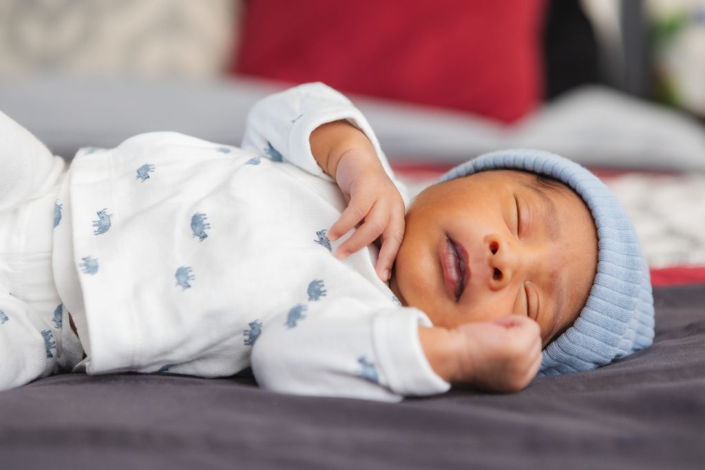 A newborn baby in a blue hat is peacefully laying on a bed in their family home.
