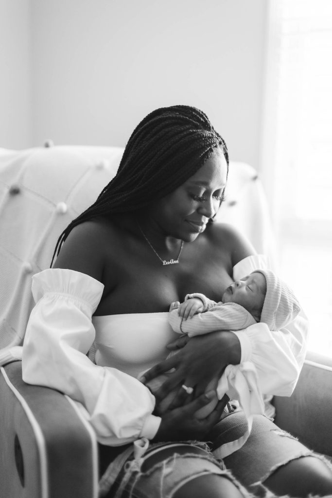 A black and white photo of a woman holding a newborn baby in a chair at their family home.