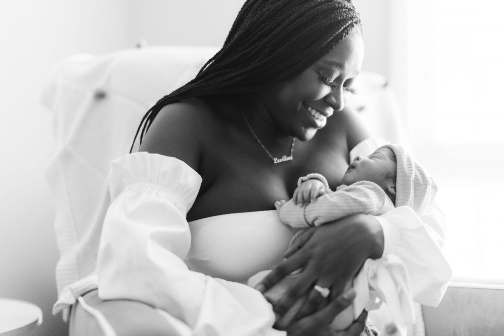 A black and white portrait of a woman holding a newborn baby in their family home.