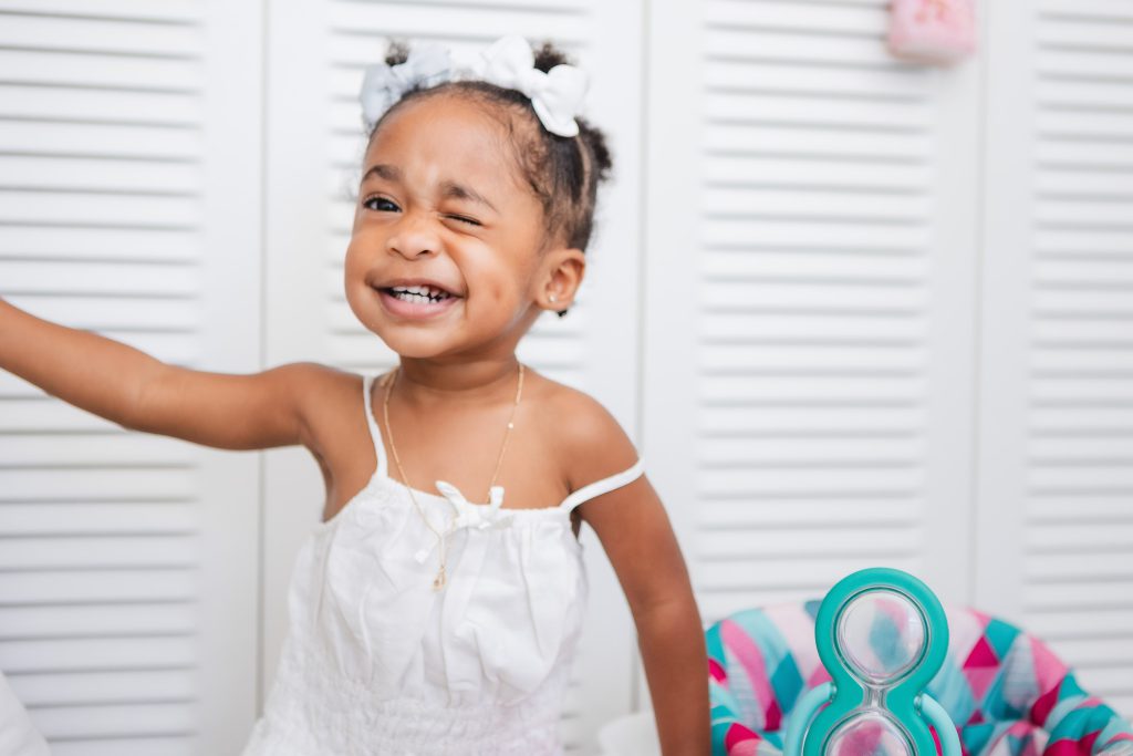 A newborn baby in a white dress standing in front of a white dresser in their family home.