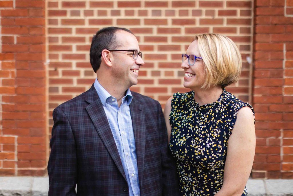 A man and woman are happily smiling in front of a brick wall while Downtown Annapolis serves as a picturesque backdrop for their family portraits.