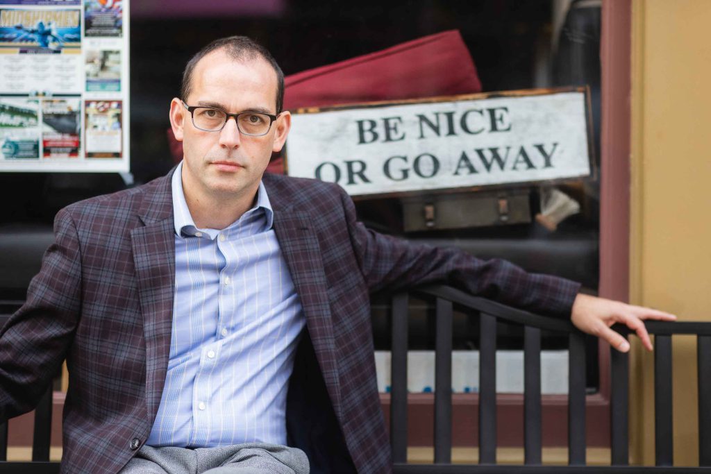 A man sitting on a bench with a sign that says be nice or go away in downtown Annapolis.